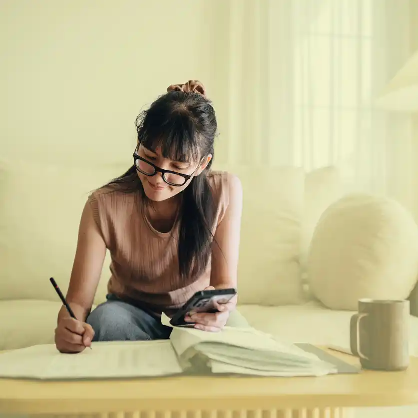 Modern woman with glasses and phone in hand writing into booklet.