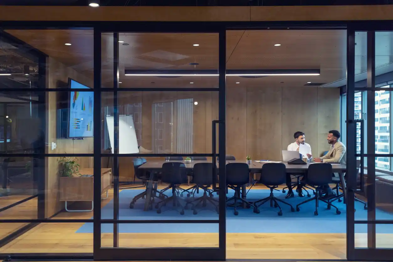 Two financial copywriters in business setting discussing across boardroom table behind glass doors.